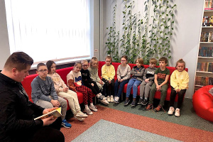 Uczniowie siedzący na czerwonej kanapie. Po lewej stronie chłopiec w czarnej koszuli czytjący dzieciom książkę. W tle białe regały z książkami, kwiaty i okna